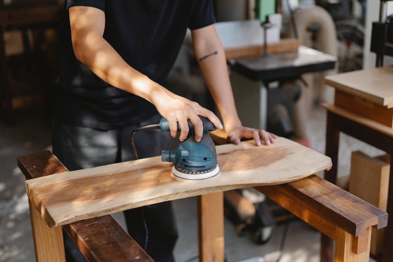 about-us-02 A craftsman uses a power sander on a wooden plank in a well-lit workshop.